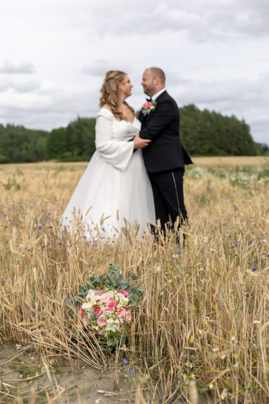 Brudbukett ligger på en åker med ett brudpar i bakgrunden efter lantligt bröllop på Vikbolandet, porträttfotograf Anna Esmelöv